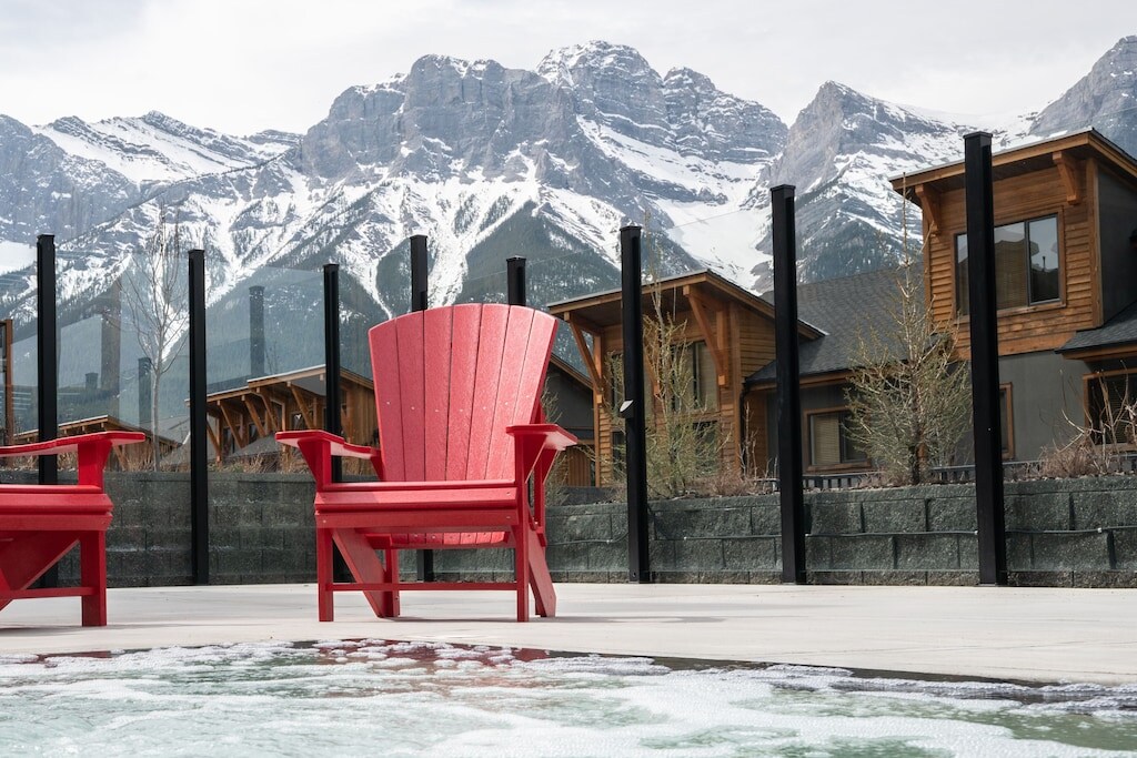 Two red Adirondack chairs are positioned by a hot tub, overlooking a scenic view of snow-capped mountains. The surrounding area is enclosed with glass panels, and wooden buildings are visible in the background, complementing the natural landscape.