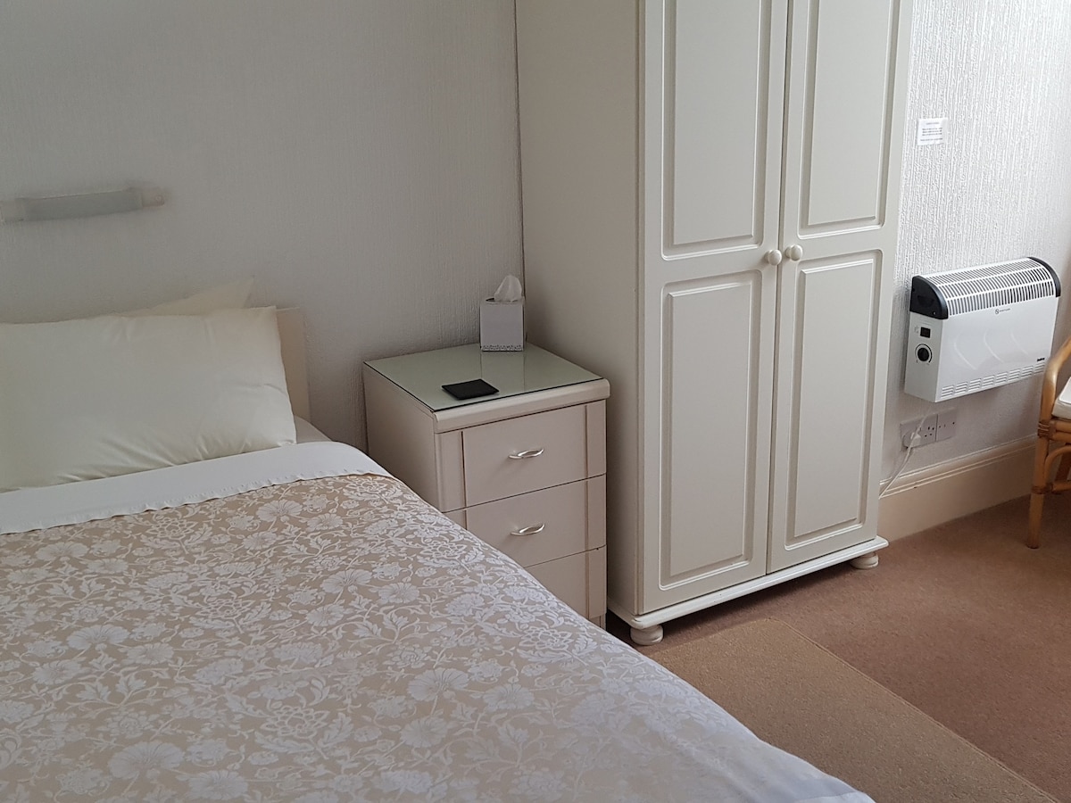 A bright sleeping area is furnished with a spacious bed featuring soft white linens. A light-colored wardrobe and bedside table create a tidy look, while a small armchair is positioned nearby. Natural light fills the room, highlighting the neutral color palette.