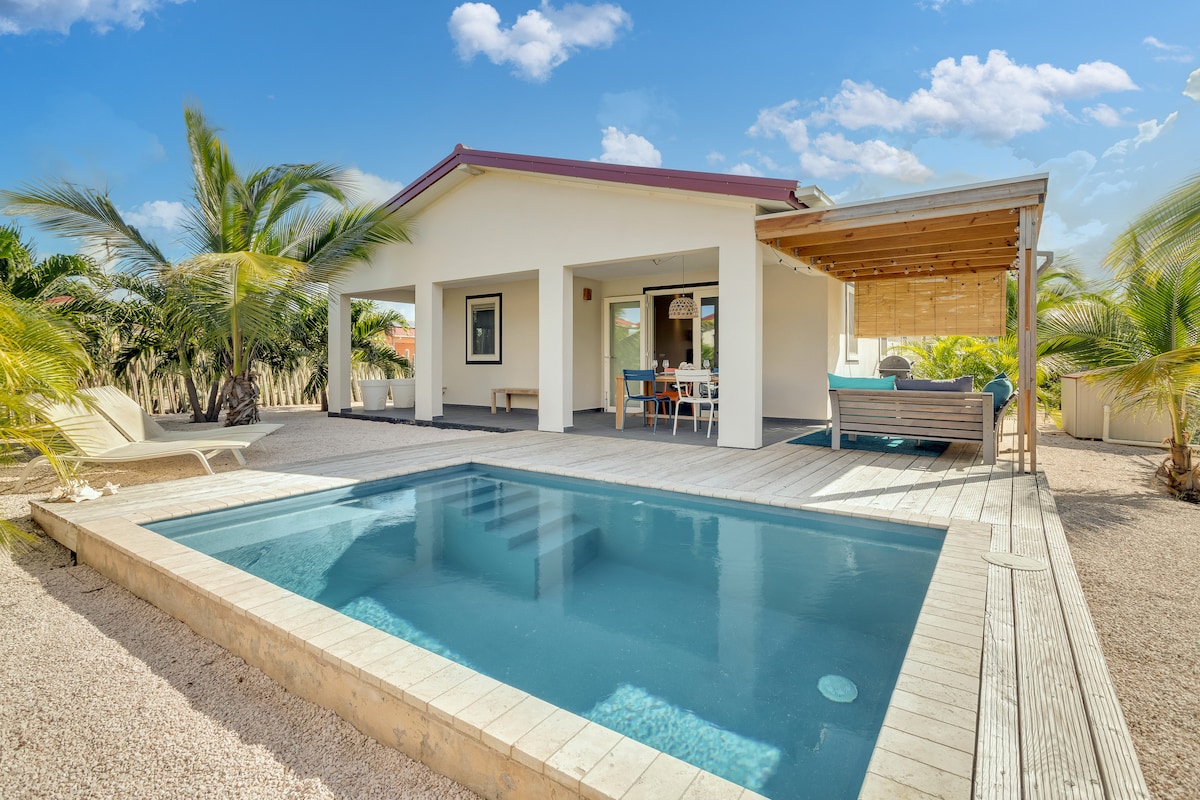 A modern villa features a small private swimming pool surrounded by tropical landscaping. The covered patio area includes seating for six under a wooden roof, offering shade. Large glass doors open to the living space, enhancing indoor-outdoor flow, with sun loungers positioned by the pool.