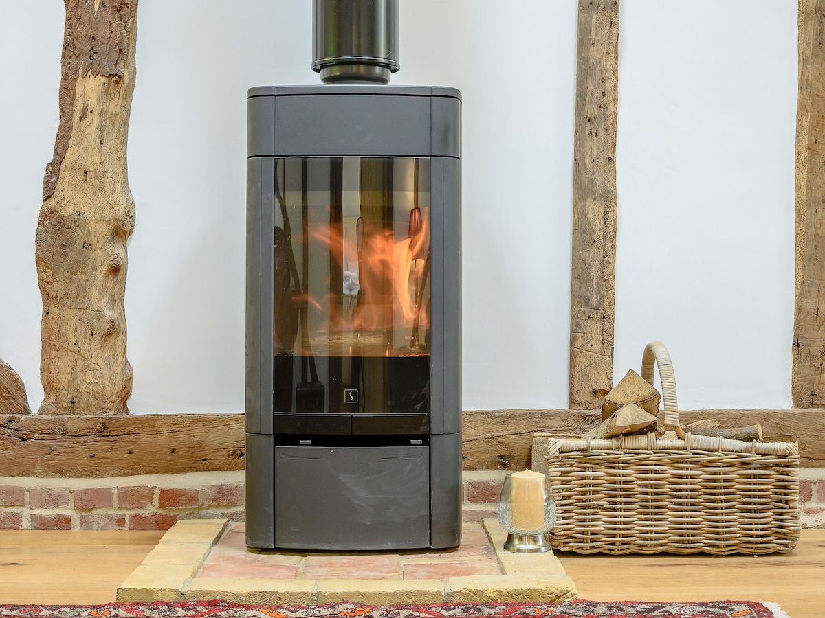A modern wood burner is positioned against a rustic feature wall with exposed wooden beams. Flames can be seen dancing inside, providing a gentle glow. A wicker basket filled with logs sits beside the burner on a stone hearth, enhancing the cozy ambiance.