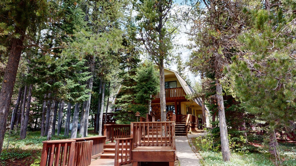 A path leads to the entryway of the cabin, surrounded by tall trees. The structure features an expansive deck with multiple seating areas, inviting relaxation in a natural setting. Sunlight filters through the leaves, creating a serene atmosphere around the home.