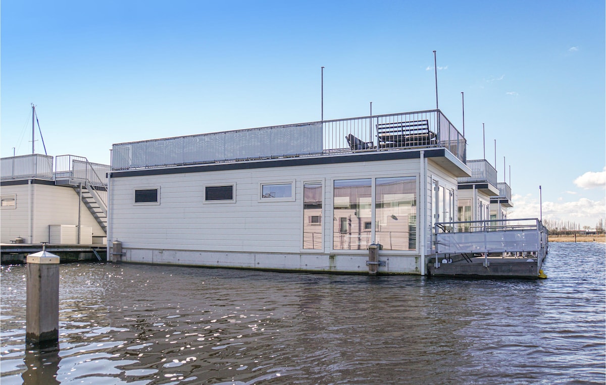 The houseboat features a spacious roof terrace, providing panoramic views of the Westeinderplassen. Large windows illuminate the exterior, with a modern design that enhances the waterfront setting. The structure is surrounded by water, reflecting the serene marina environment.