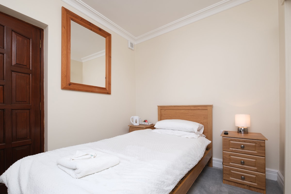 A simple and inviting bedroom features a single bed made with white linens. A nightstand with drawers sits beside the bed, accompanied by a lamp. A wall-mounted mirror enhances the lightness of the space. A kettle and cups are placed on a small table.