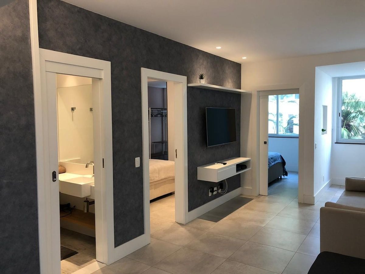 The living area features a neutral-toned wall and a mounted television, complemented by various doorways leading to functional spaces. Soft lighting accentuates the clean lines and minimal design elements, while a window allows natural light to brighten the area.