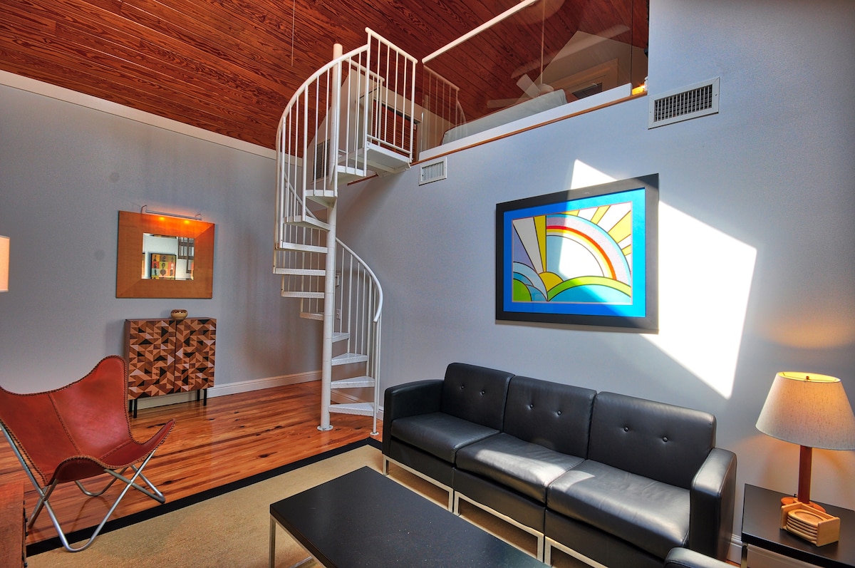 A cozy living room features a dark leather sofa and a unique, modern art piece on the wall. A spiral staircase with white railings leads to a loft area. Natural wood beams and a warm light enhance the inviting space.