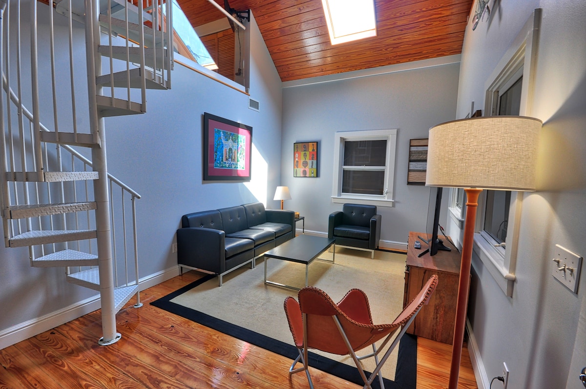 A spiral staircase ascends to a loft area in a well-lit living room, featuring high ceilings and a skylight. Two sofas are arranged around a coffee table on a neutral rug, with a lamp providing soft illumination. Colorful artwork is displayed on the walls.