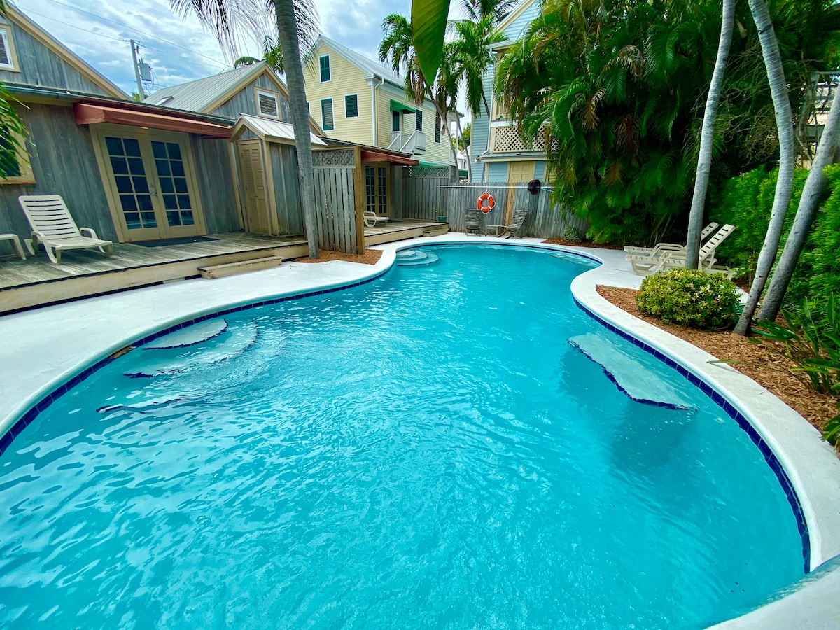 A kidney-shaped pool glistens under a partly cloudy sky, surrounded by lush tropical greenery. The pool features built-in seating areas and is framed by a clean white border. Nearby, wooden decking provides space for lounging and enjoying the serene outdoor environment.