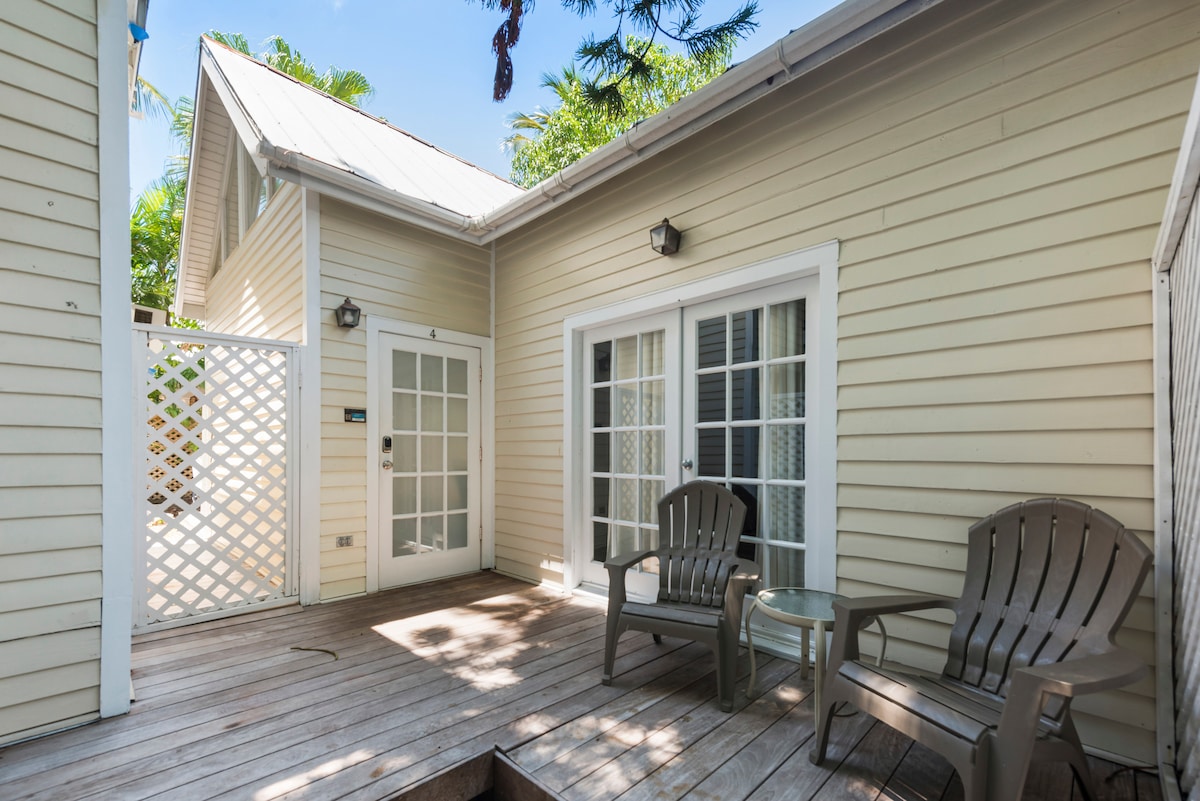 A private deck area features two gray chairs and a small glass table, surrounded by lush tropical foliage. The deck offers an inviting exterior view of the cottage, with a white lattice screen providing added privacy.