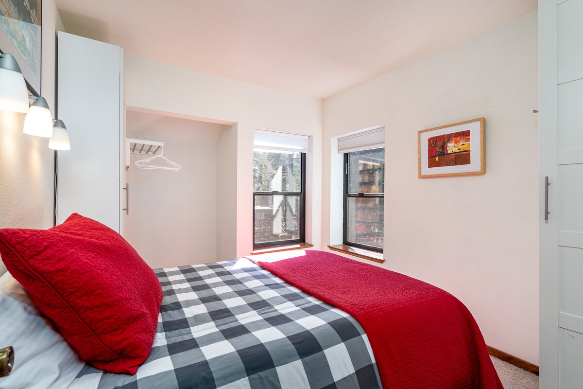 A cozy bedroom features a red and gray plaid bedding set on a spacious bed. Two windows allow natural light to illuminate the room, framed by simple white trim. A wall-mounted light fixture adds warmth, while a piece of framed artwork adds a pop of color.
