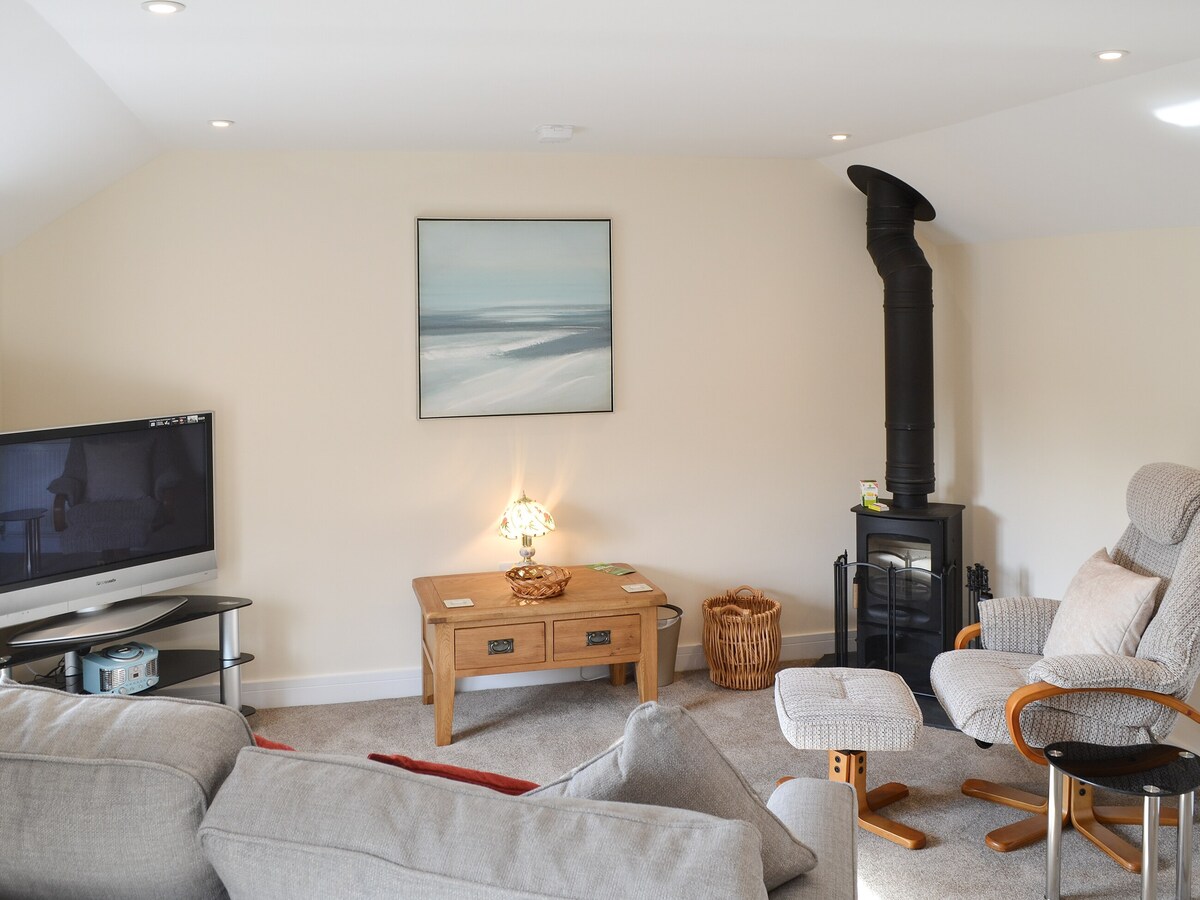 An open plan living area is presented with a light colour palette. A large sofa and a recliner accompany a coffee table, while a wall-mounted television is positioned nearby. A woodburner adds warmth, and an artwork depicting coastal scenery adorns the wall.