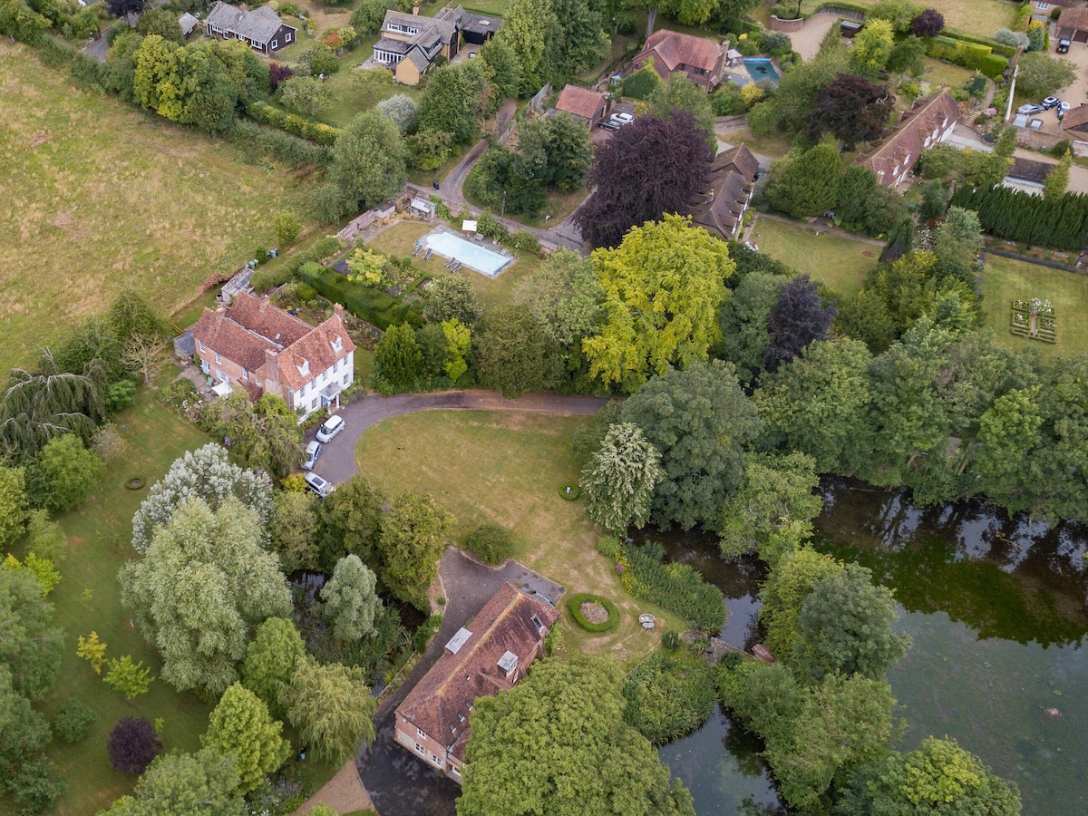 An aerial view capturing the beautiful grounds surrounding Moorhen Cottage, with lush greenery and trees bordering the landscape. The shared swimming pool is visible, along with well-kept gardens and the picturesque lake nearby. Charming cottages and homes are nestled throughout the countryside.