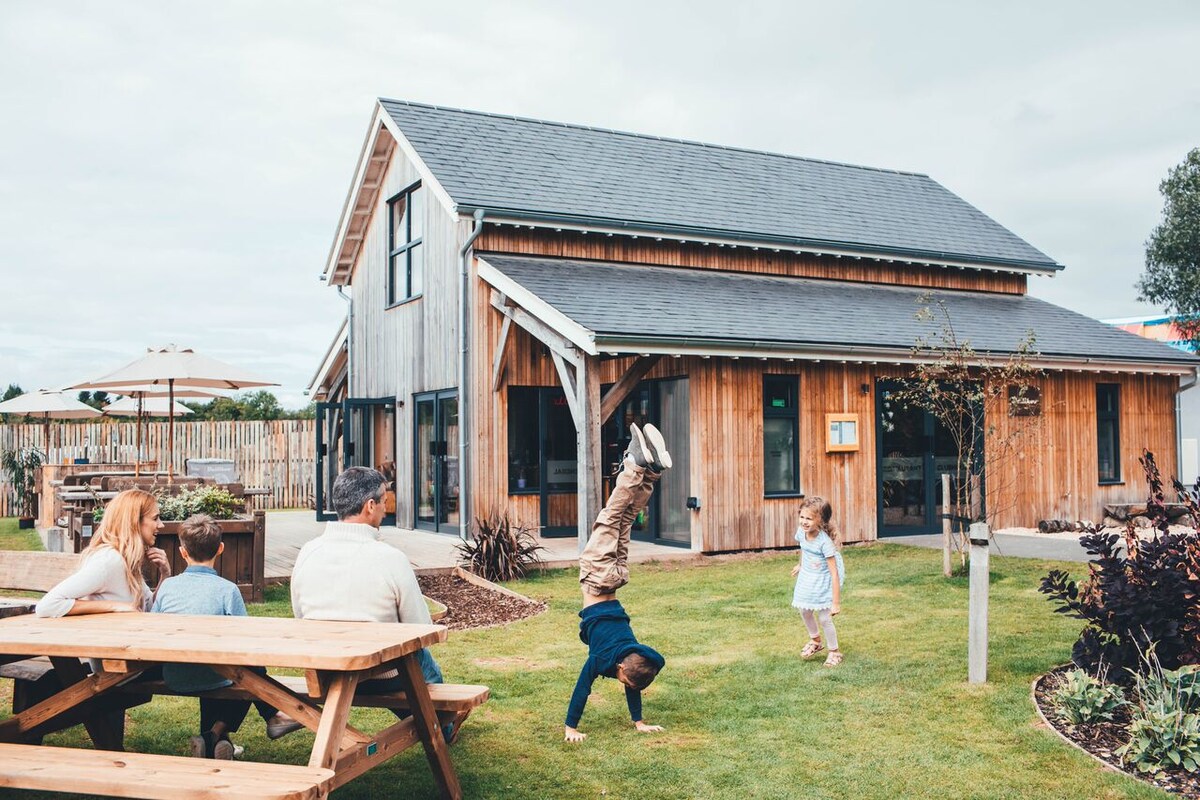 A modern timber-clad holiday home is depicted with a spacious lawn in front. Families are enjoying time outdoors, with one child performing a handstand. Picnic tables are arranged nearby, surrounded by greenery and garden features, creating an inviting atmosphere for gatherings.