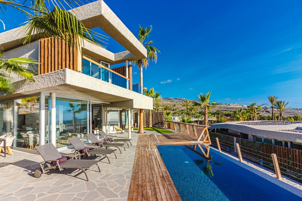 The exterior of a modern villa showcases a spacious terrace featuring sun beds and an infinity pool. Lush palm trees surround the property, and large glass doors provide views of the interior. A clear blue sky complements the contemporary design.