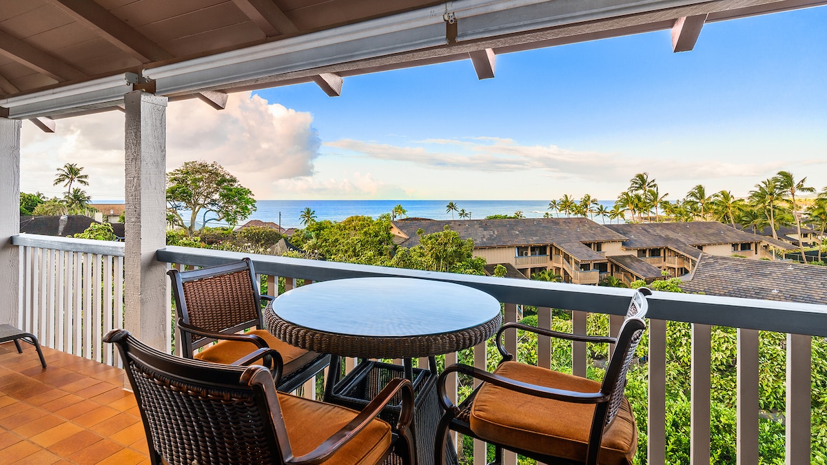 A private lanai is showcased, featuring a circular table and two chairs with cushions. Lush greenery surrounds the area, offering a view of the ocean in the distance. The sky displays soft clouds and a light blue hue, creating a relaxing outdoor space.
