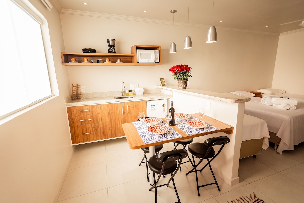 A well-appointed kitchenette features a countertop, coffee maker, microwave, and mini refrigerator. A dining table with two chairs is set for a meal, complemented by decorative tableware. In the background, a spacious bed with neatly arranged linens is visible, creating a functional and welcoming space.