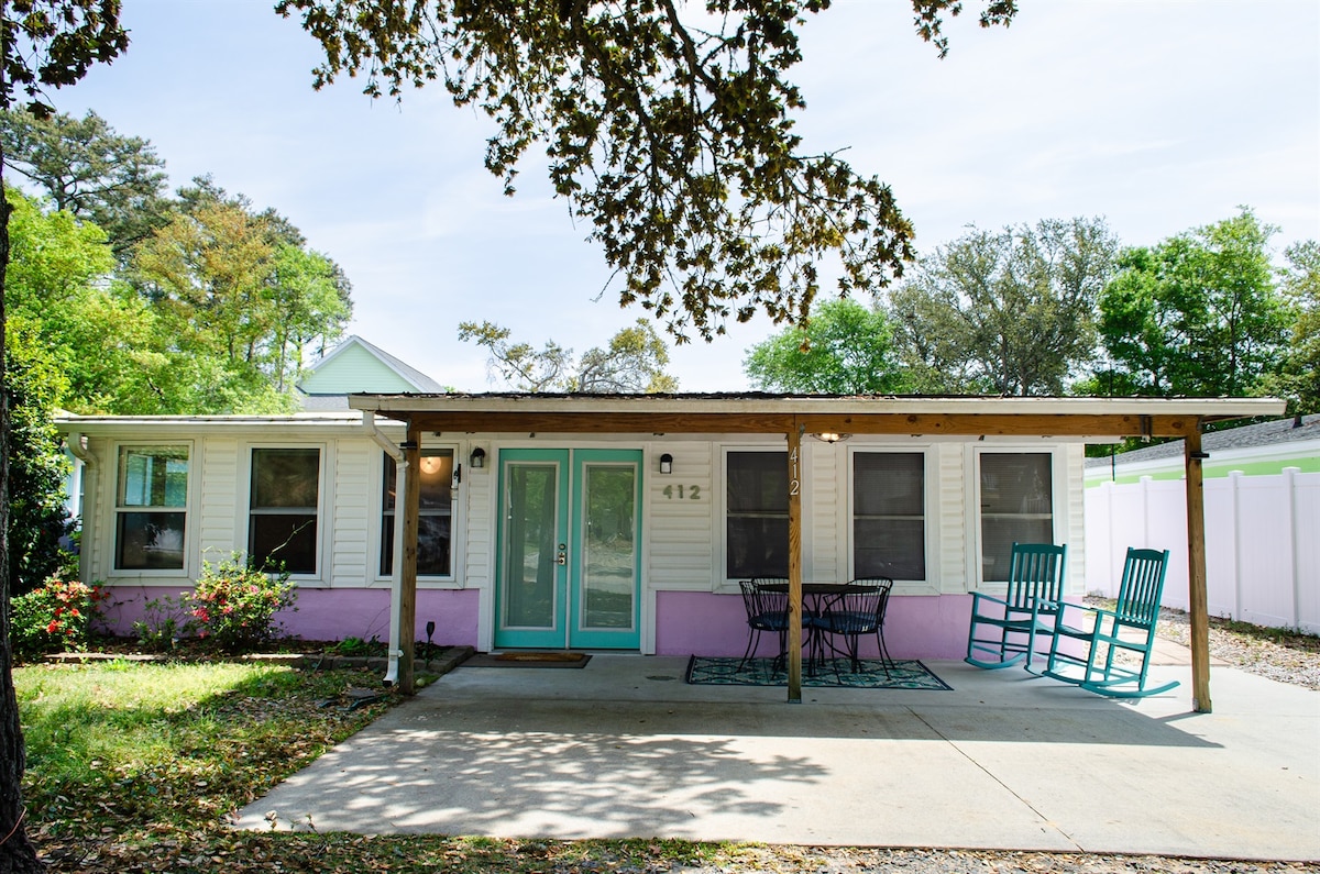 A charming cottage exterior is displayed, featuring a covered front patio with a small table and two rocking chairs. The bright teal door welcomes guests, and the pink accent below the windows adds a playful touch. Lush greenery surrounds the property.
