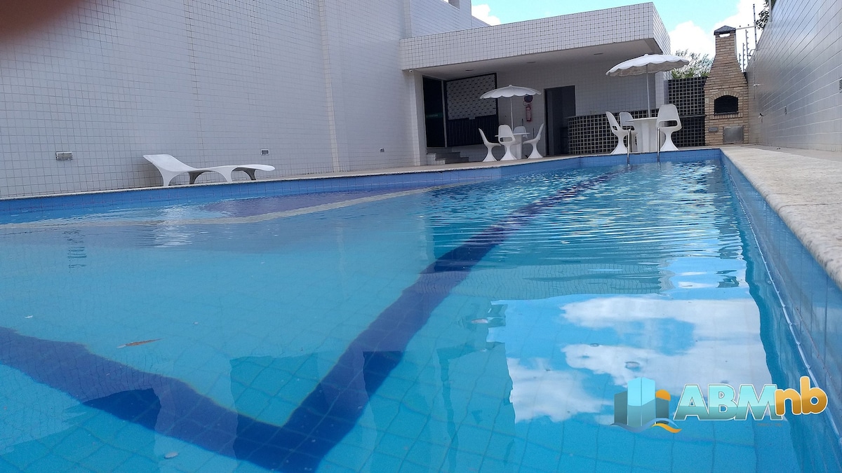 A clear blue swimming pool is featured, reflecting the sky above. Two white lounge chairs are positioned nearby, alongside a shaded seating area under an umbrella. A barbecue grill is visible in the background, enhancing the outdoor space.