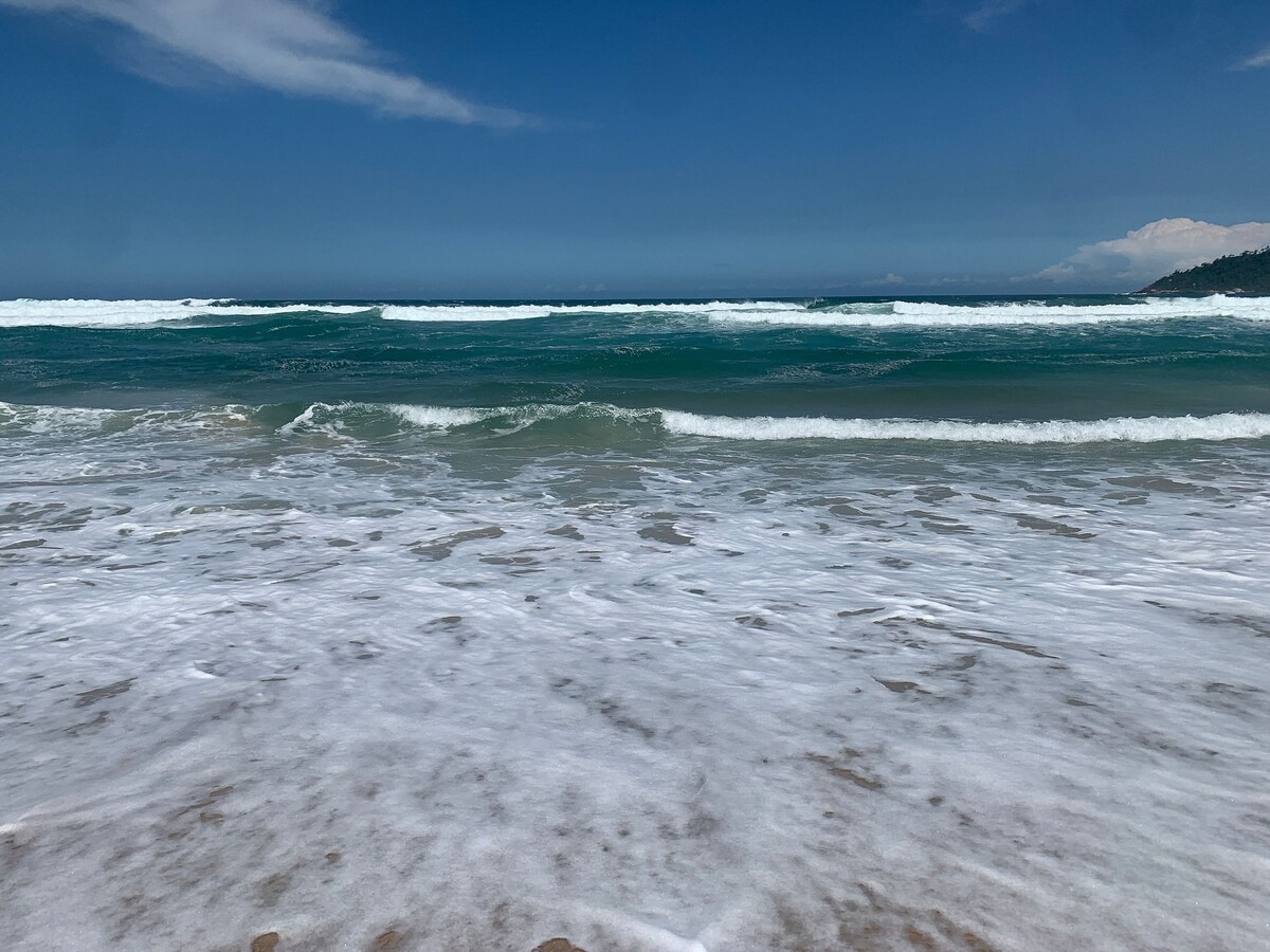 Gentle waves roll onto the sandy shore, with clear blue skies above. The horizon blends with serene ocean waters, showcasing varying shades of blue and green. White foam from the waves creates a soft contrast against the sandy beach, inviting a peaceful coastal atmosphere.