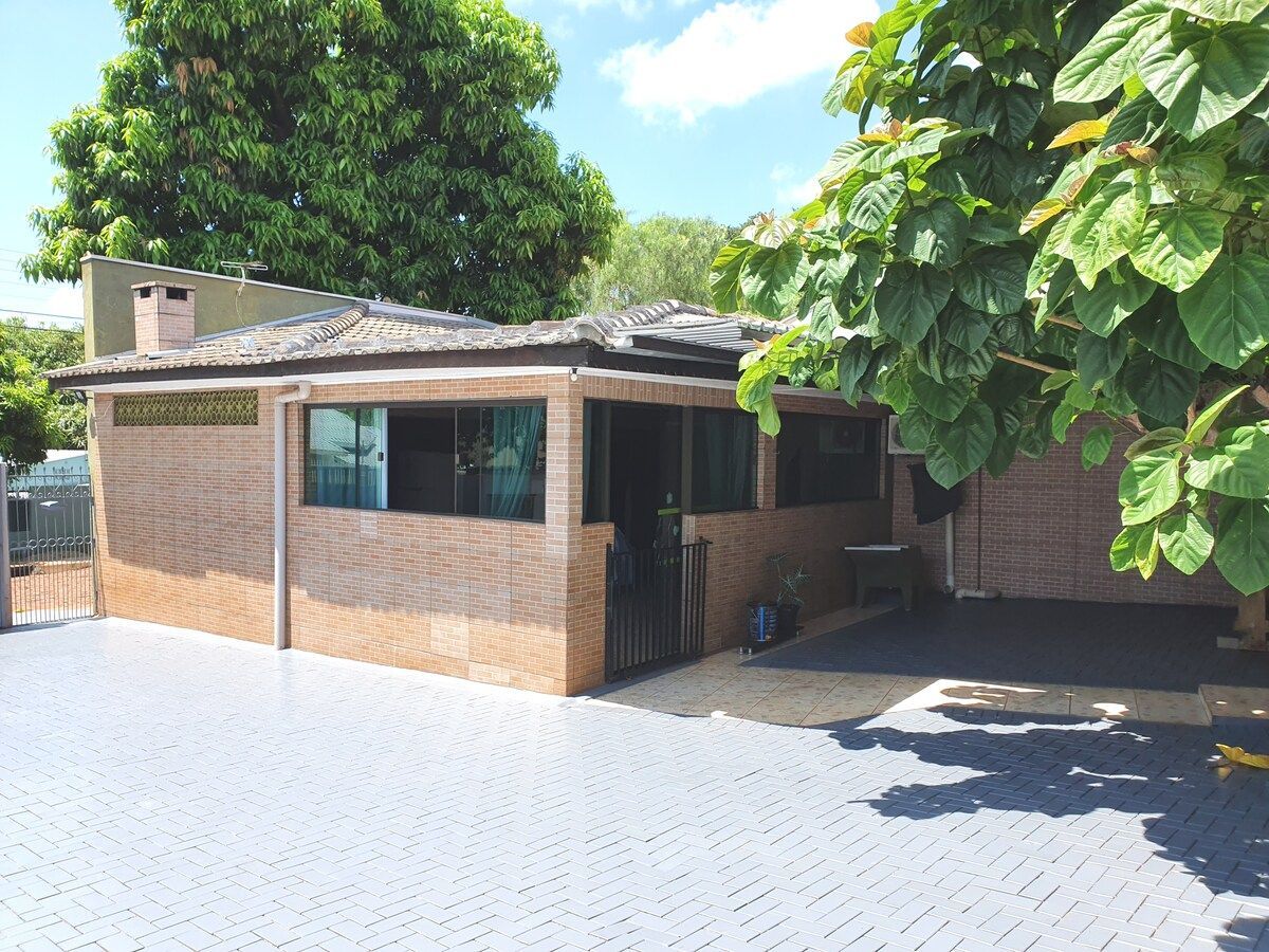 A spacious exterior area showcases a brick house surrounded by greenery. Large windows offer natural light and views of the outdoor space, which features a paved surface and a tree providing shade. A fence adds privacy and security to the property.