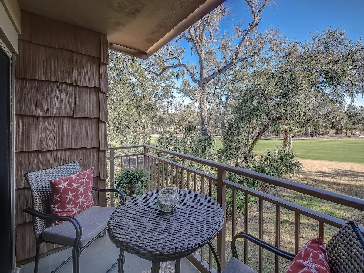 A private balcony is showcased with a round table and two chairs, ideal for outdoor relaxation. The view includes lush greenery and hole 9 of the Atlantic Dunes golf course, framed by trees that provide a serene backdrop.