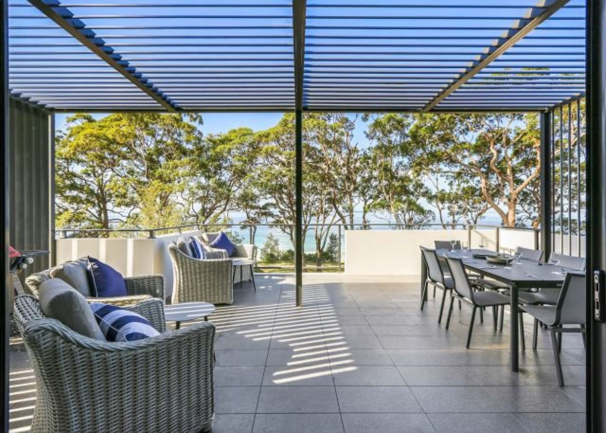 An outdoor terrace features a spacious dining table and several lounge chairs. Shaded by a slatted pergola, the area offers scenic views of trees and the water in the distance, creating an inviting space for relaxation and alfresco dining.