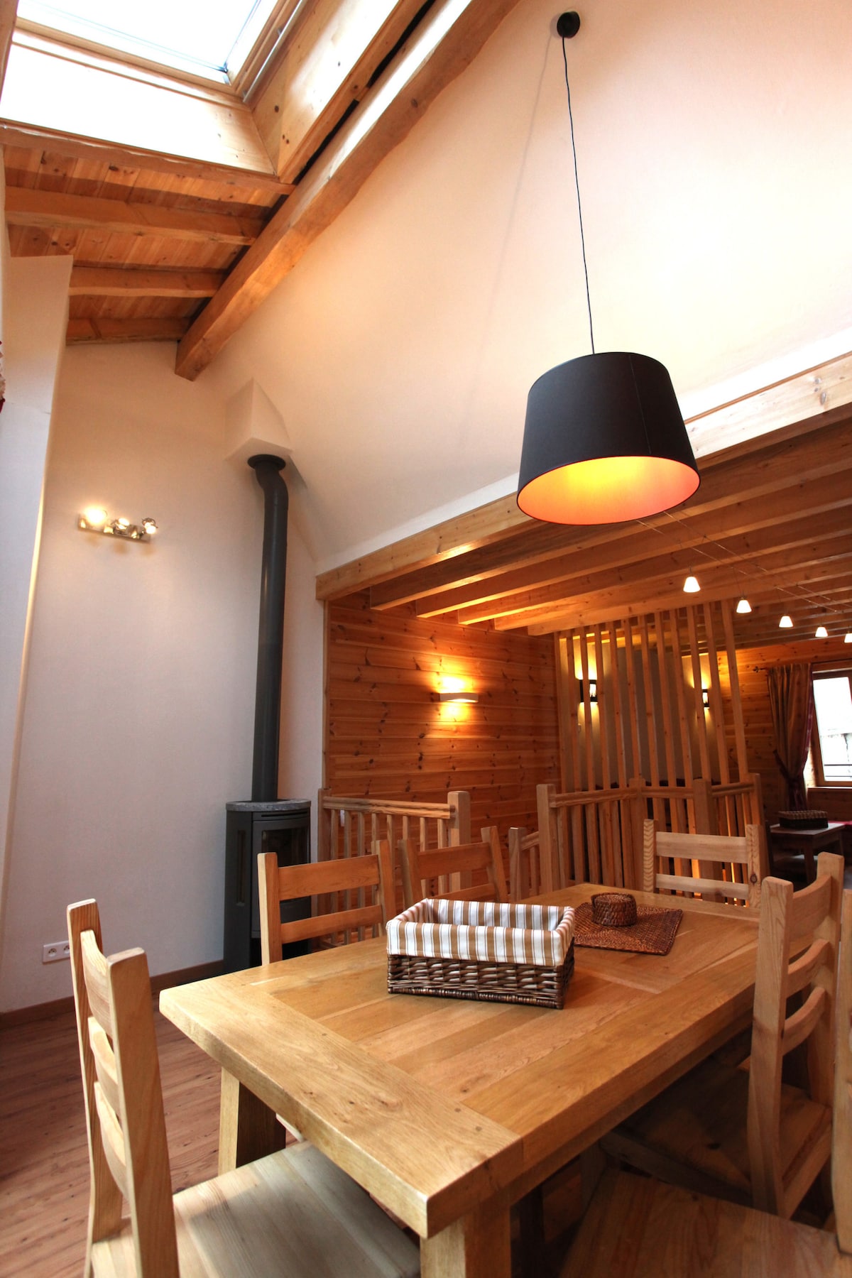 The dining area features a wooden table surrounded by six chairs, illuminated by a pendant light. A wood stove is positioned in the corner, and wooden beams highlight the sloped ceiling. Natural light is provided by a skylight, creating a warm environment.