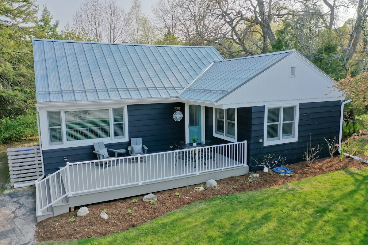 The exterior of the Howard House is showcased, featuring a modern design with dark siding and a metal roof. A welcoming front porch includes two chairs for relaxing, while a grassy area highlights landscaped greenery. Surrounding trees add a touch of nature to the serene setting.