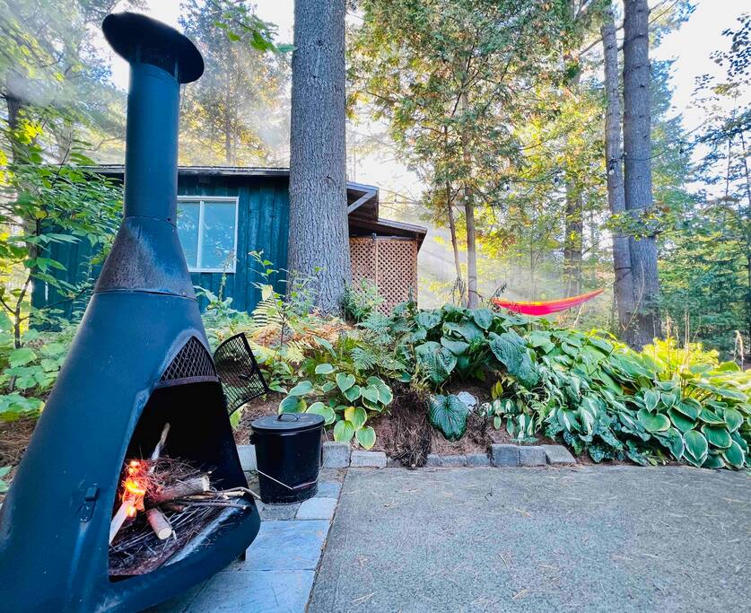 An outdoor area features a black chiminea with logs prepared for a fire. Lush green plants and a vibrant red hammock create a serene backdrop, while the forested setting adds to the tranquil atmosphere. The wooden cabin can be seen in the background.