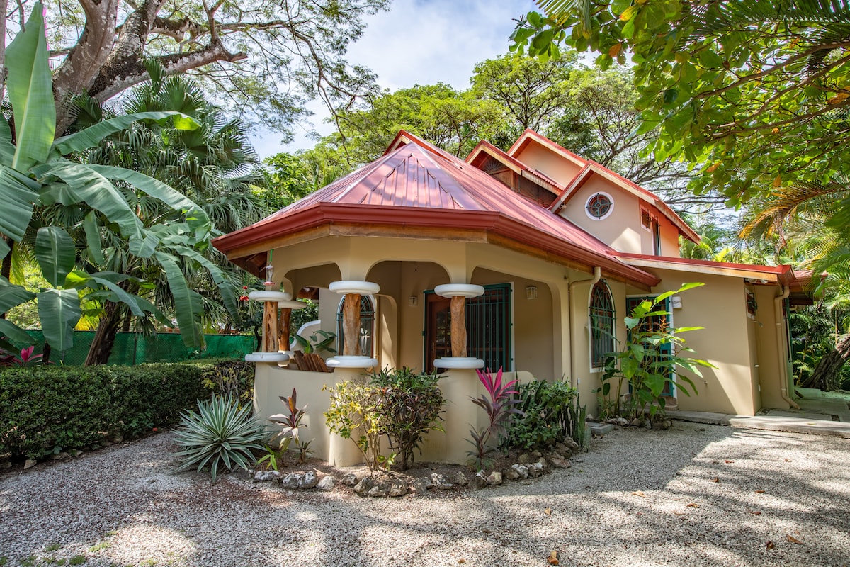 The exterior of the home is presented, featuring a charming design with a red metal roof and decorative arches. Lush greenery surrounds the property, including tropical plants and trees. A gravel path leads up to the entrance, emphasizing a tranquil, natural setting.