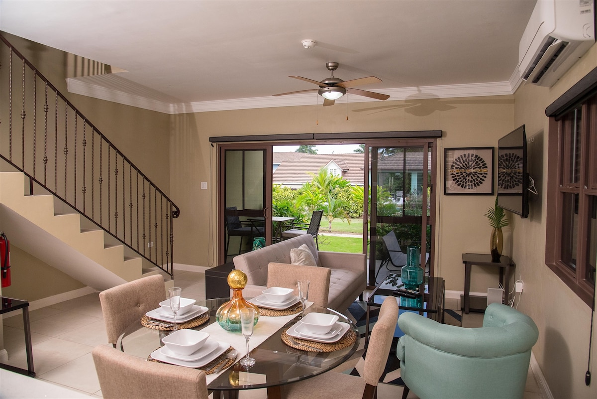 An open-concept living area features a dining table set for four and a plush sofa. Natural light enters through large glass doors, providing views of the manicured landscape outside. A ceiling fan circulates above, enhancing the comfortable ambiance of the space.