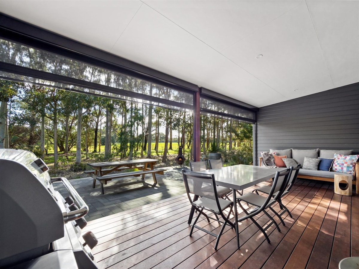 A shaded outdoor deck features a large dining table surrounded by black folding chairs. A gas BBQ is positioned nearby, with a cozy seating area equipped with colorful cushions. Lush trees can be seen in the backdrop, enhancing the tranquil atmosphere.