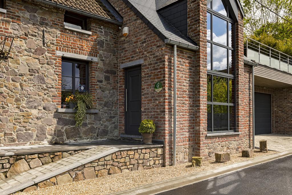 The exterior of a renovated stone farmhouse is presented, featuring a blend of large windows and textured brickwork. A small potted plant adorns the entrance, while a pathway made of small stones leads to the door. A garage is visible in the background.