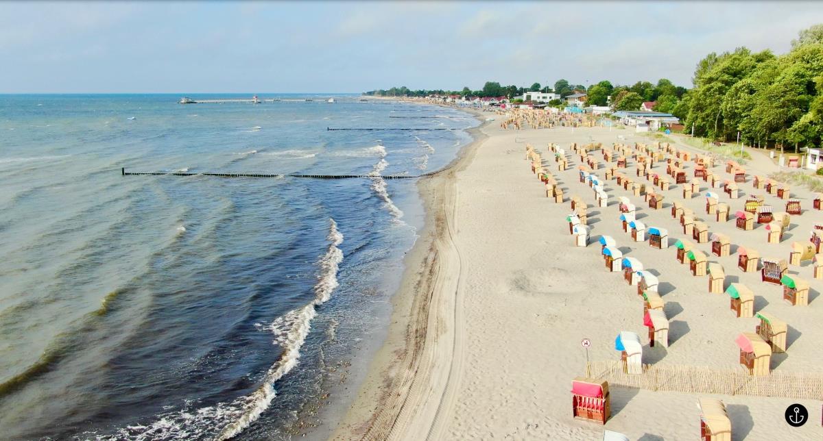 A sandy beach is seen, lined with colorful beach chairs scattered across the shore. Gentle waves roll in, meeting a smooth sandy expanse that stretches toward the horizon. In the background, greenery and a coastal community are visible, adding to the scenic beach environment.
