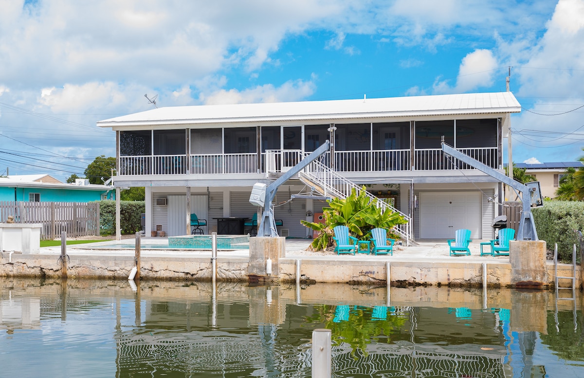 The stilted home is surrounded by water, showcasing a spacious screened porch, and outdoor seating. A new pool is visible in the backyard alongside the canal. A staircase leads to the upper level, and the property features a neat, well-maintained exterior under a bright sky.
