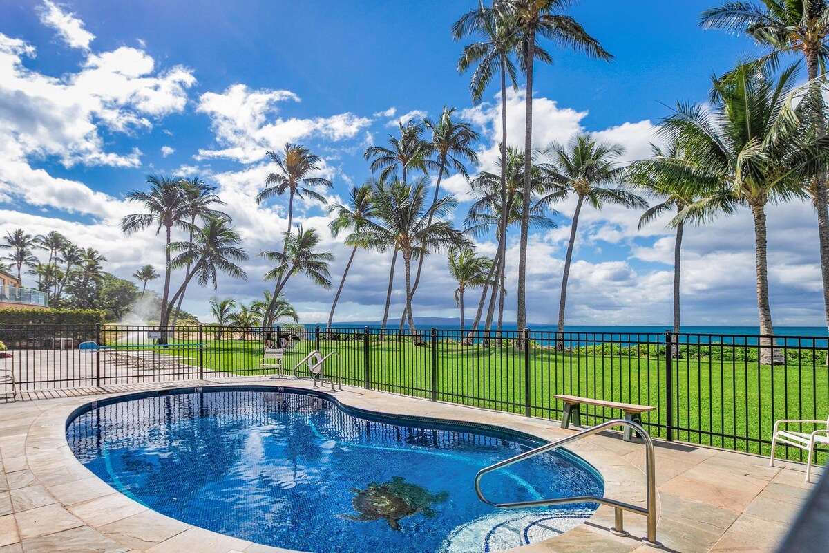 A gated swimming pool surrounded by lounge chairs offers an inviting space for relaxation. Lush green grass and tall palm trees frame the pool area, with stunning ocean views in the background under a clear blue sky with scattered clouds.