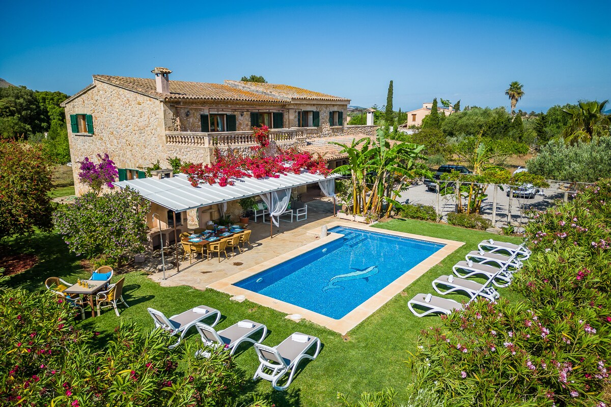 A rural dwelling is set amidst lush greenery, featuring a swimming pool surrounded by sun loungers. Colorful flowers adorn the outdoor area, while shaded terraces offer seating for relaxation. The house's balcony overlooks the pool and gardens, providing an inviting outdoor space.