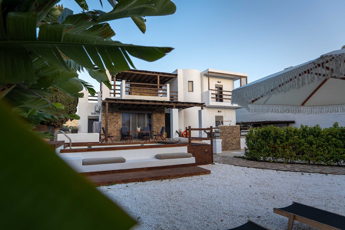 The exterior of the villa is showcased, featuring a two-level structure with wooden balconies and stone accents. A spacious outdoor area is visible, complete with a white stone patio and lounge chairs surrounded by greenery. The overall ambiance reflects a serene and inviting setting.