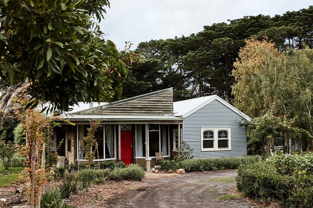 The cottage exterior showcases a blend of rustic styles with a metal roof and wooden accents. A bright red front door is framed by tall windows, while a landscaped garden with various plants and trees surrounds the entrance, providing a welcoming view.