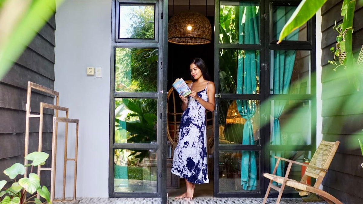 A guest stands at the entrance of the villa, holding a book. The entryway is framed by large glass doors, which open to a view of the lush garden. A cozy chair is positioned beside the entrance, and vibrant green curtains enhance the welcoming atmosphere.