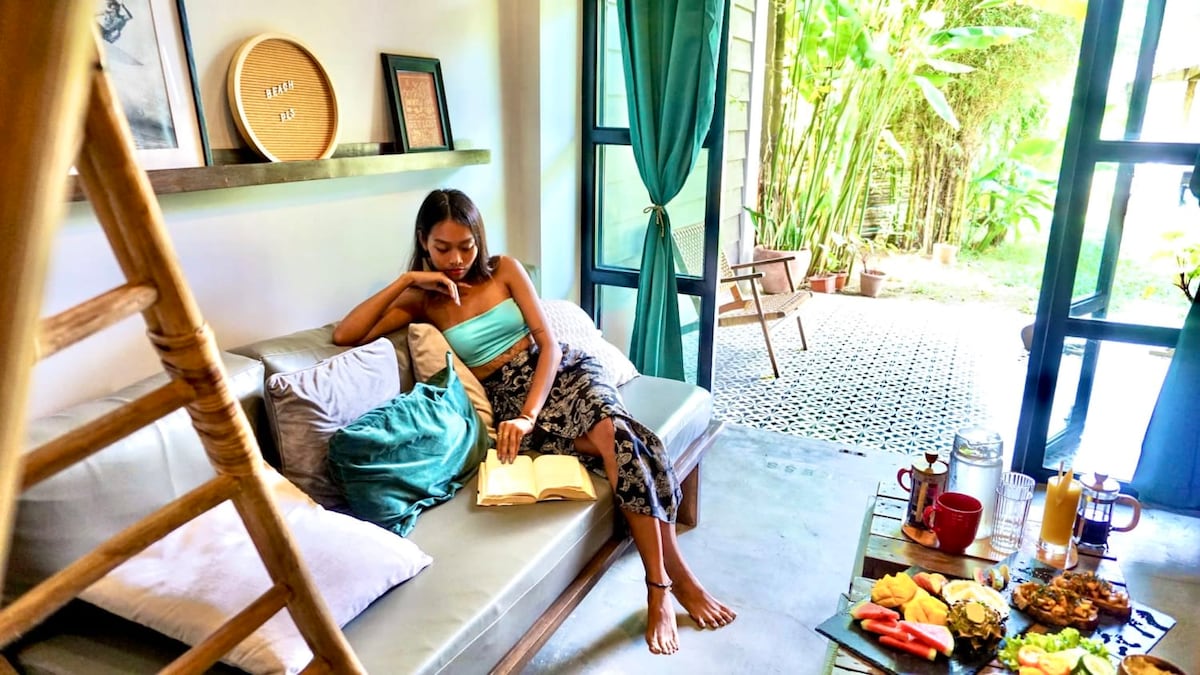 A comfortable sitting area features a light-colored sofa adorned with soft pillows, set against a backdrop of lush greenery visible through large glass doors. A table is arranged with breakfast items, highlighting a welcoming space for relaxation and enjoyment.