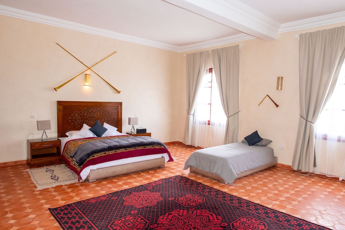 A spacious bedroom is furnished with a king-sized bed dressed in rich linens and a single bed with a simple cover. Two large windows allow natural light to fill the room, while a decorative rug adds color to the tiled floor. Elegant curtains frame the windows.