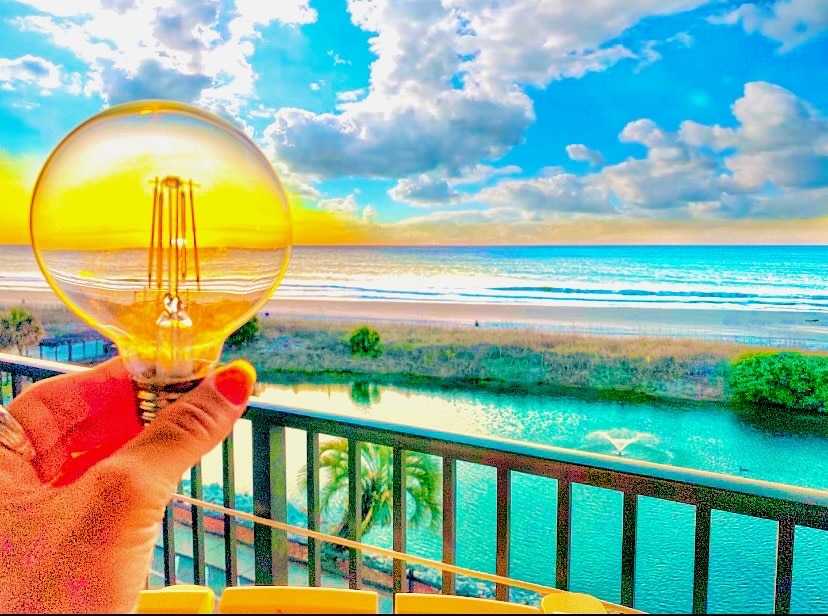 A hand holds a large light bulb against a vibrant sunset sky, showcasing the golden sun reflecting over the ocean. Below, the pond is visible with greenery, adding to the serene oceanfront atmosphere. The balcony railing is partially seen in the foreground.