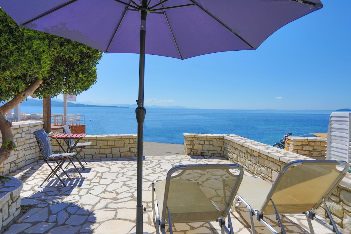 A sunlit terrace overlooks a clear blue sea, featuring two lounge chairs and a table shaded by a large purple umbrella. The stone-paved area is bordered by lush greenery, creating a serene outdoor space with expansive water views.