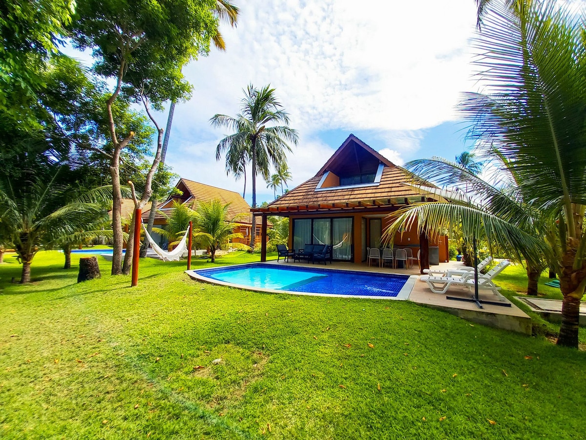 An inviting bungalow set amidst lush tropical landscaping features a private pool and is surrounded by palm trees. A spacious outdoor area includes two sun loungers near the pool and a hammock draped between two trees, offering a tranquil retreat.