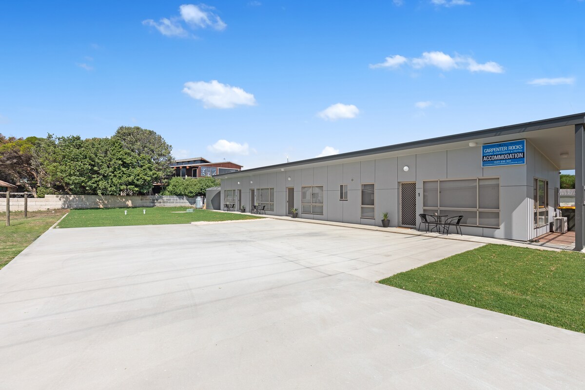 A modern motel-style complex is shown, featuring a spacious concrete driveway and lawn area. Outdoor seating is available near the entrance, with clear blue skies in the background and lush greenery surrounding the property.