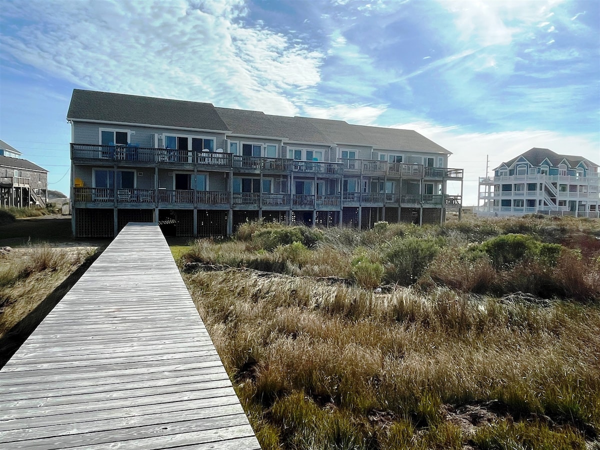 The exterior view of the Sea Whisper Condos is presented, showcasing a multi-unit building with balconies. A wooden walkway leads towards the building, surrounded by natural grasses and brush, under a clear blue sky with scattered clouds.