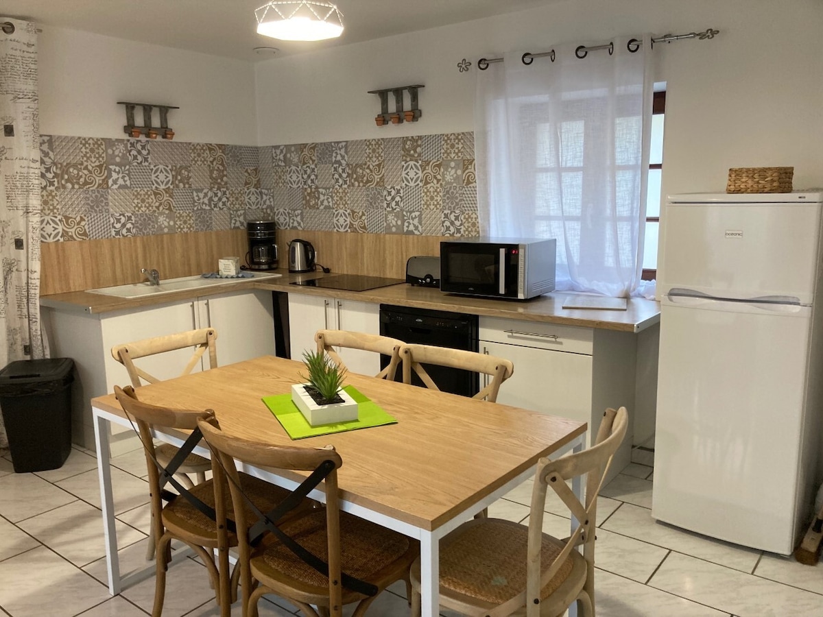 A modern kitchen is equipped with essential appliances, including a refrigerator, microwave, and dishwasher. A wooden dining table with six chairs occupies the center, complemented by decorative elements such as a small plant. Natural light filters through a window adorned with a sheer curtain.