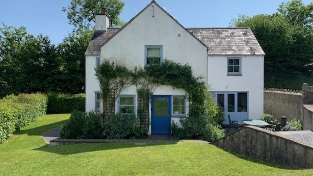 A charming two-story cottage is set within a green landscape, featuring a white exterior with blue accents. Lush vegetation adorns the front, while a patio area is visible with outdoor furniture. The surrounding garden offers a spacious lawn for relaxation.