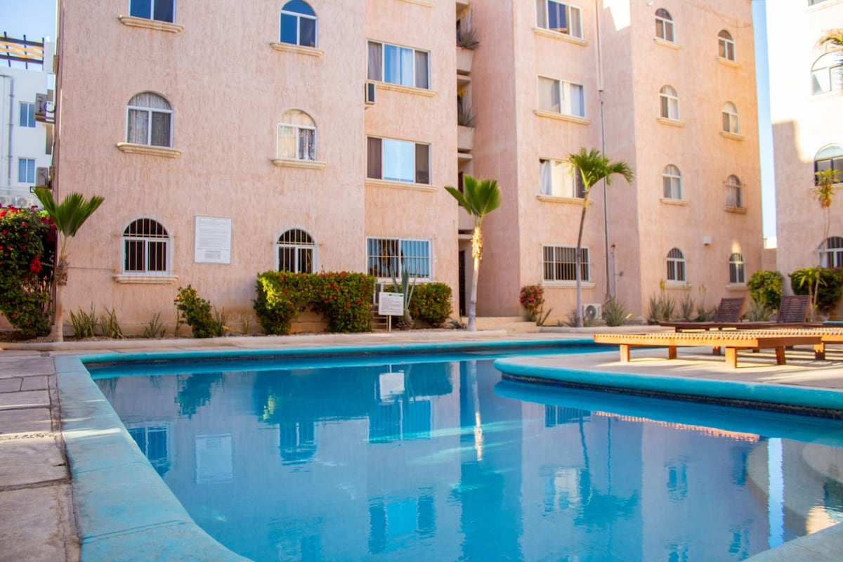 A tranquil pool area is surrounded by a well-maintained courtyard, featuring a clear blue swimming pool reflecting the beige facade of the surrounding building. Palm trees and vibrant greenery enhance the serene outdoor space, inviting relaxation.