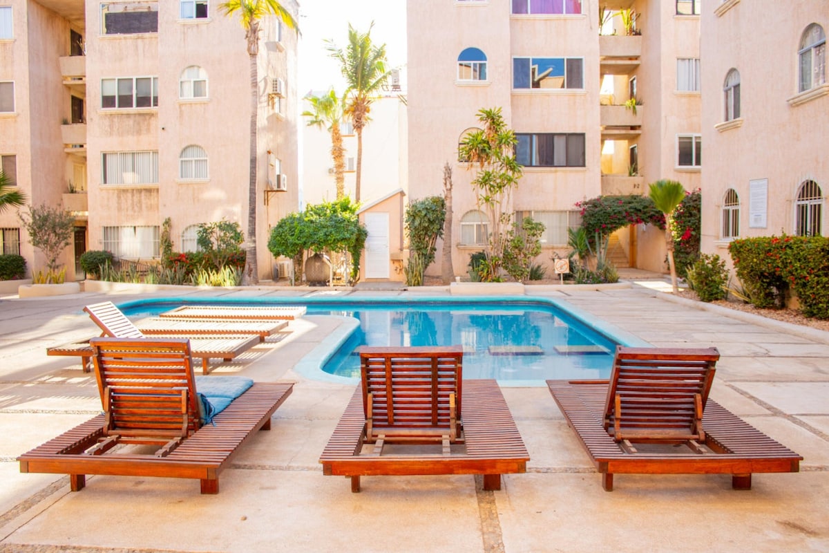 The image showcases a common pool area surrounded by neatly maintained greenery. Three wooden lounging chairs are positioned beside the pool, inviting relaxation. The building's façade features light-colored walls and large windows, adding to the serene atmosphere of the space.